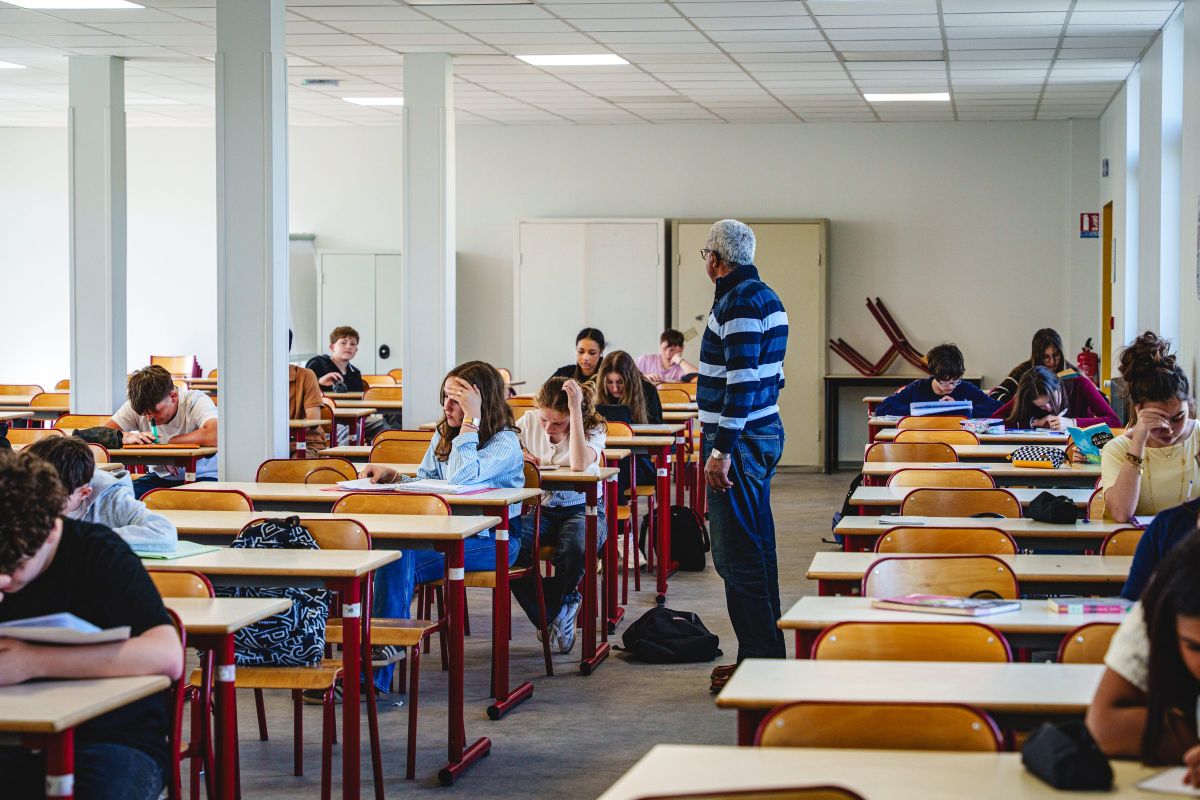 Salle d'examen au collège avec des élèves penchés sur leur copie et un surveillant